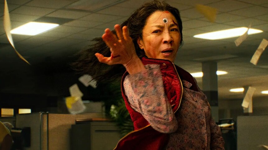 Michelle Yeoh wearing a red vest and a googly eye on her head. Her hand is up in a martial arts position
