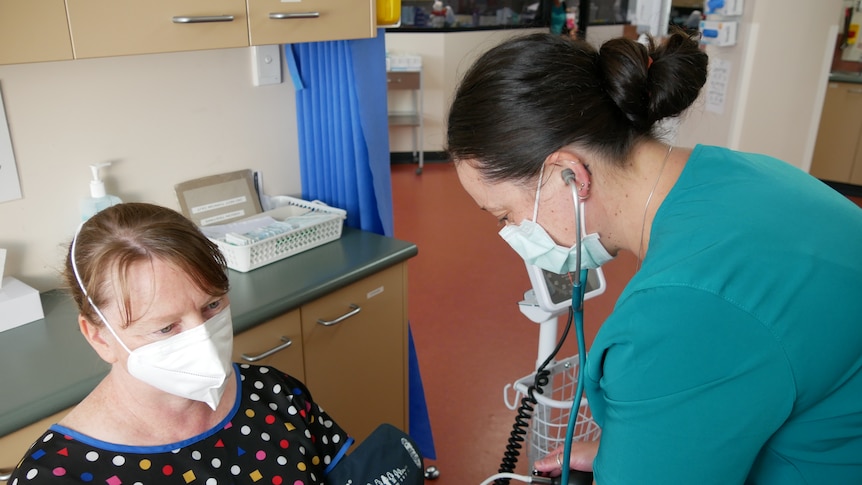 A woman in scrubs and a facemask leans over another woman wearing a facemask checking blood pressure.