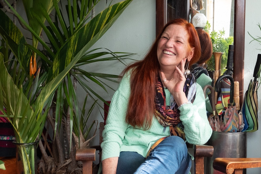 A woman with long red hair sits in a chair in her home, smiling.