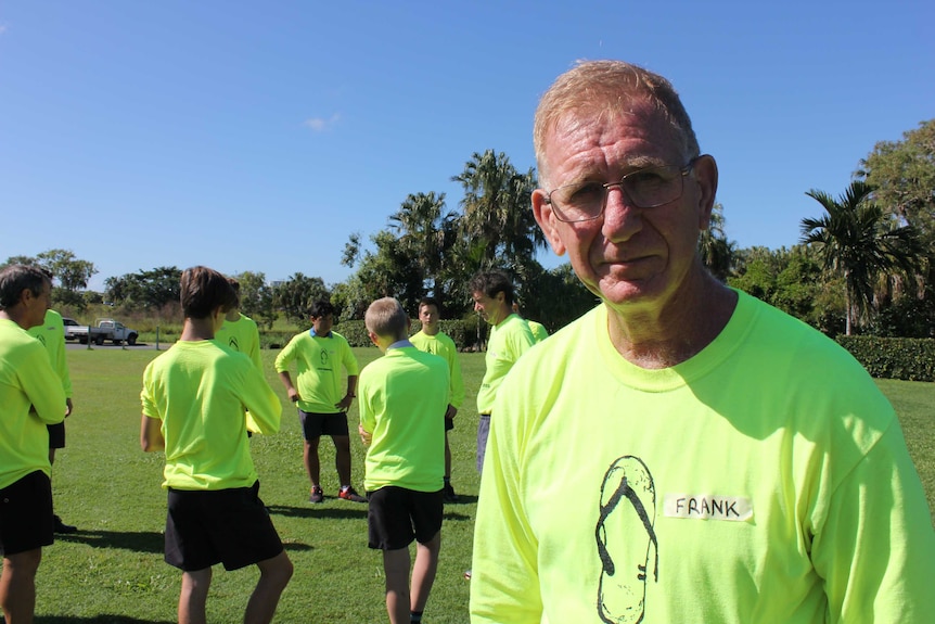 A man dressed in fluoro yellow with a broken thong on his shirt smiles as a group of teenage boys completes a class outside