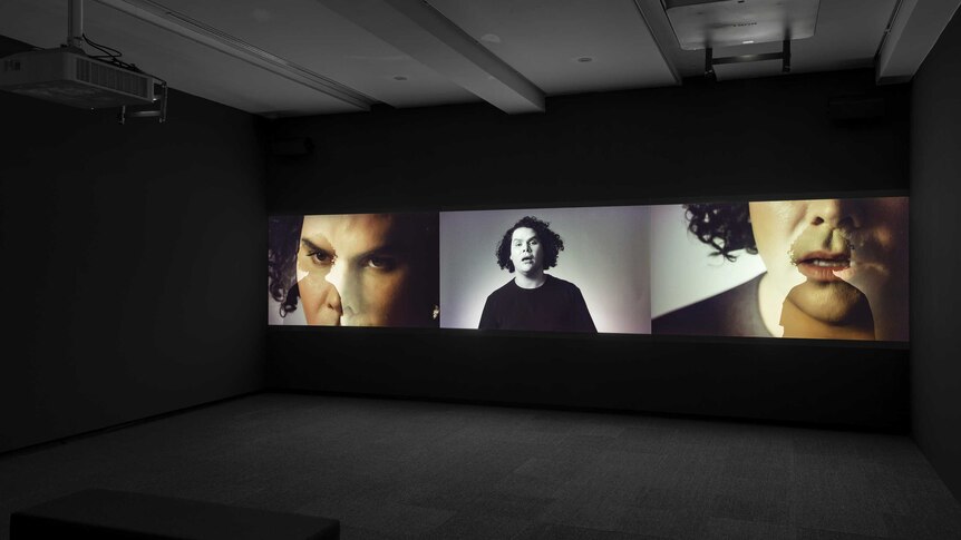 In a darkened cinema style room, a three panel video work displays the same artist (Christian Thompson) from a variety of angles