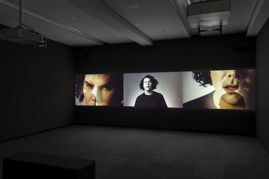 In a darkened cinema style room, a three panel video work displays the same artist (Christian Thompson) from a variety of angles