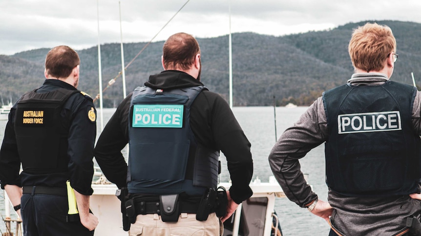Three men wearing vests that say POLICE stand on a jetty looking into a boat.