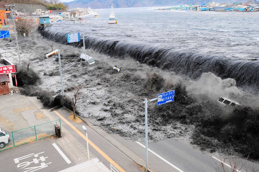 A massive wave of dirty brown water spills onto a road, washing away cars and street signs.