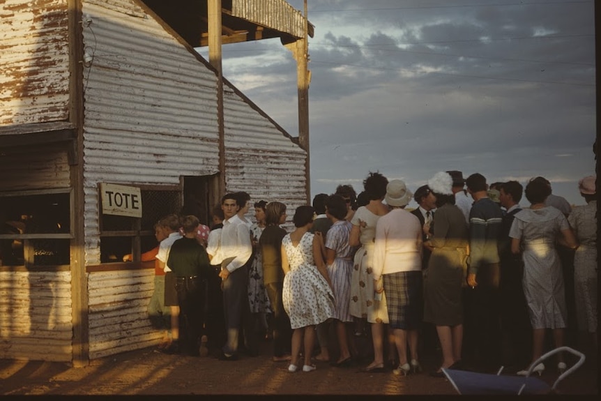 People in early 1960s fashion line up to bet at a counter in a tin shed with a sign that says "tote".