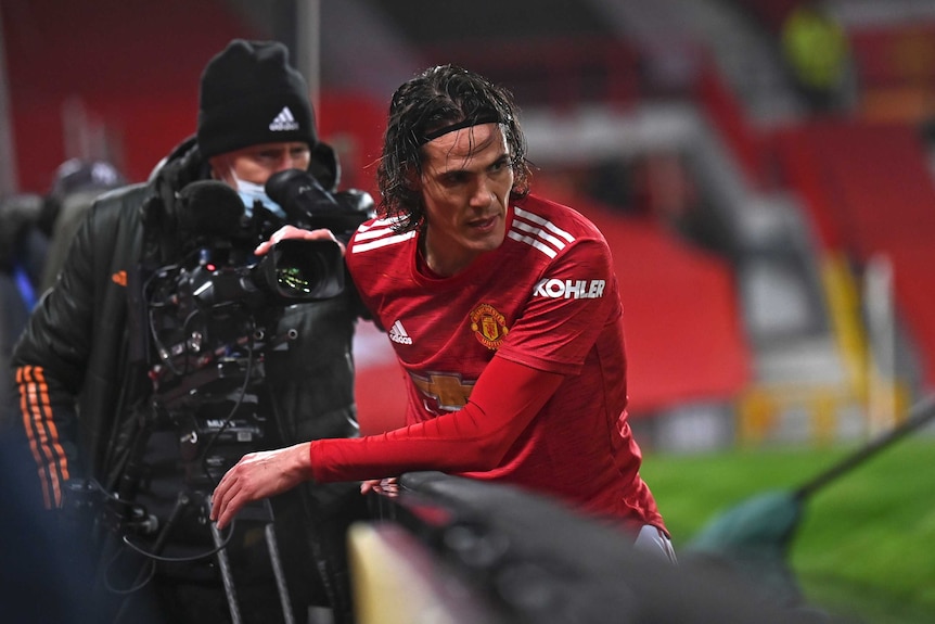 A player in a red Manchester United jersey leans over an advertising hoarding during a game.