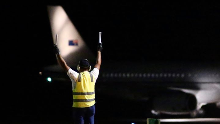 A Christmas Island airport ground staff member holds two batons in the air at night with a plane in the background.