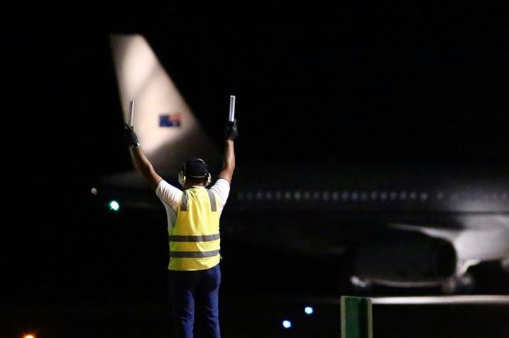 A Christmas Island airport ground staff member holds two batons in the air at night with a plane in the background.