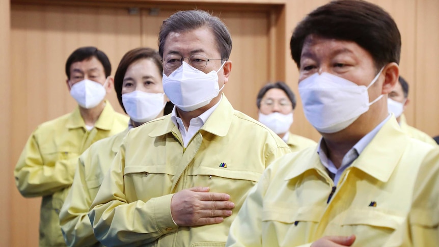 A group of people wearing masks and yellow raincoats raise their right hands to their chest.