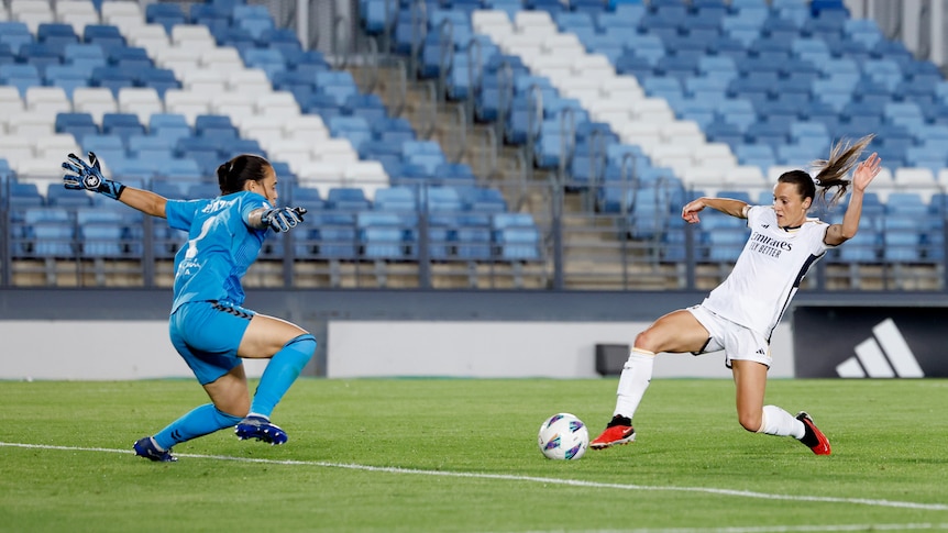 A soccer player wearing white pokes the ball with her foot at another player wearing blue during a game
