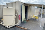 A cream-coloured shipping container with a door opening onto a covered concrete patio. A water tank is attached to one end.