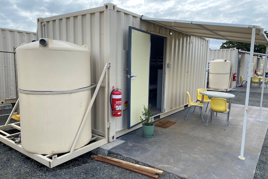 A cream-coloured shipping container with a door opening onto a covered concrete patio.  A water tank is attached to one end.