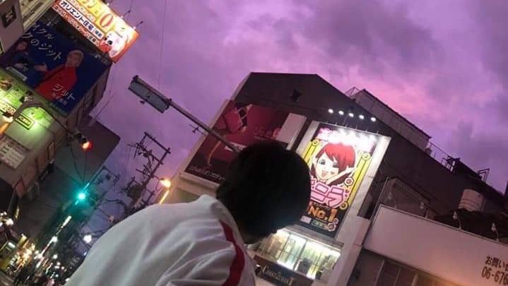 A man on a street filled with colourful  signs in Japanese looks up at vivid purple clouds,