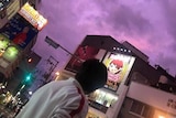 A man on a street filled with colourful  signs in Japanese looks up at vivid purple clouds,