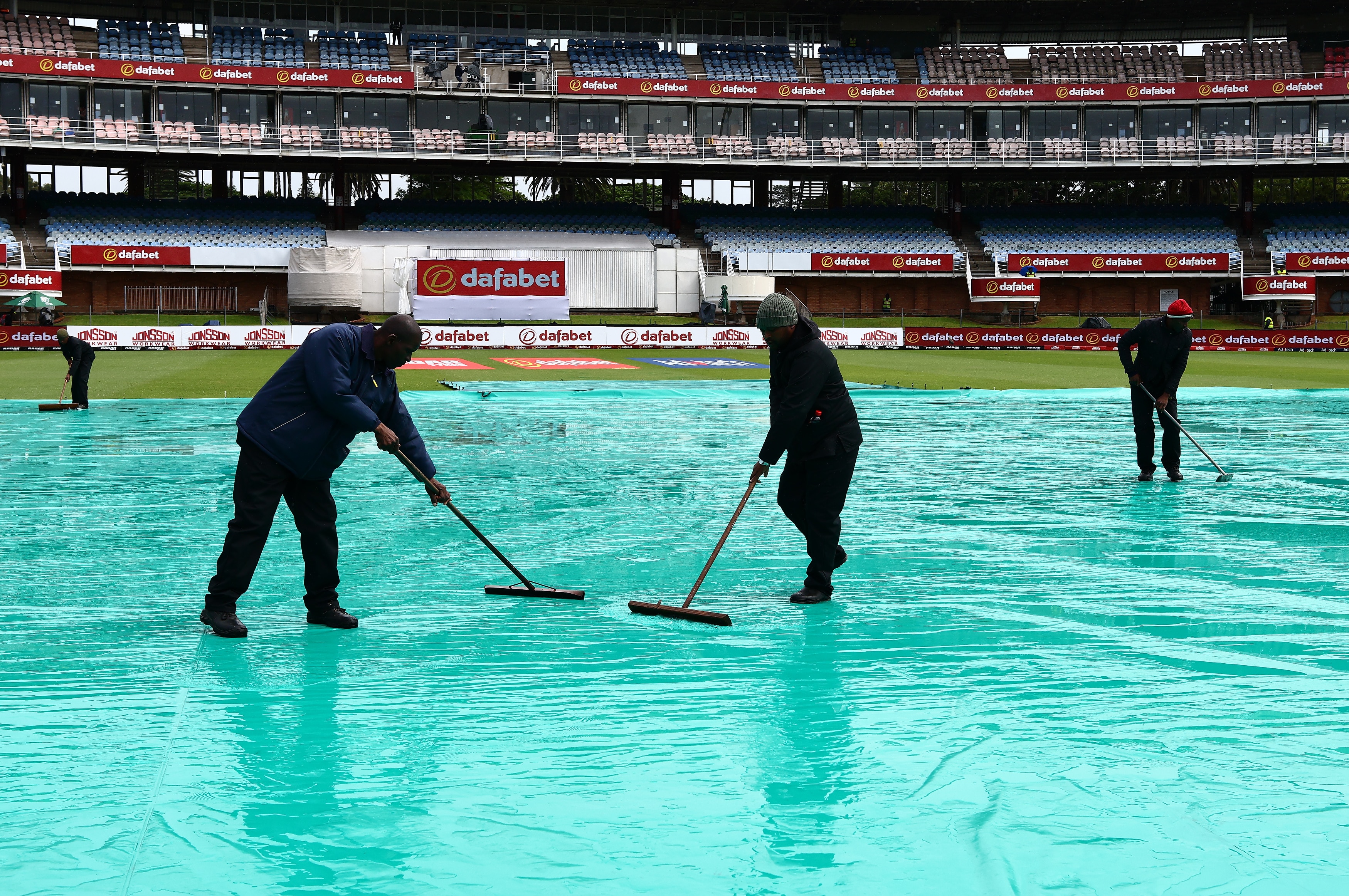 Three men in black rake water off covers in the middle of a cricket field.
