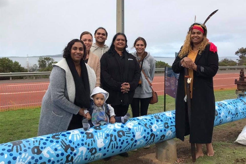 A group of Aboriginal people and an SA Water spokesperson stand beside a blue pipe with black and white hands painted onto it.