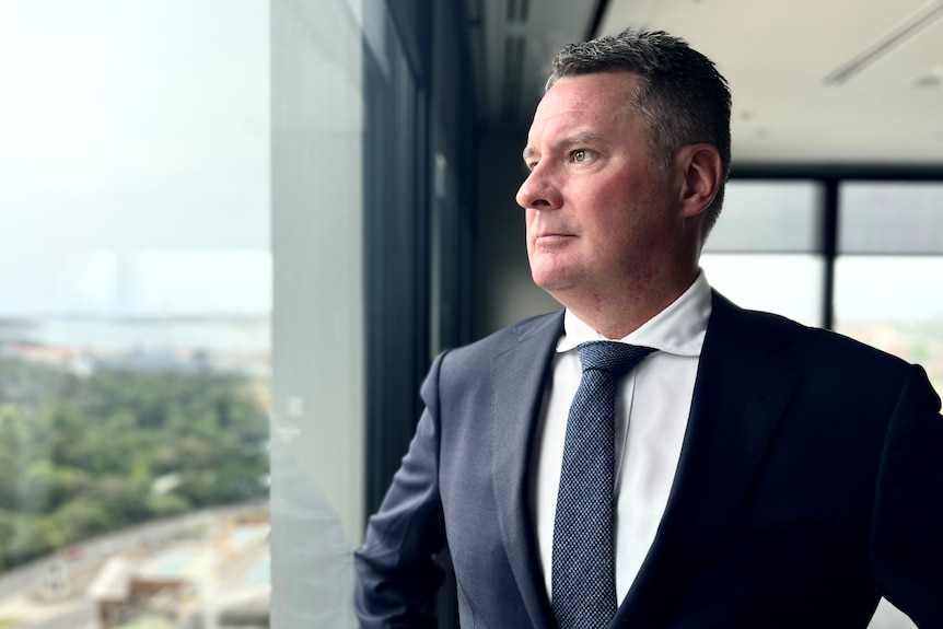 A man waring a dark blue suit and tie stands with his hands on his hips as he looks out the window.