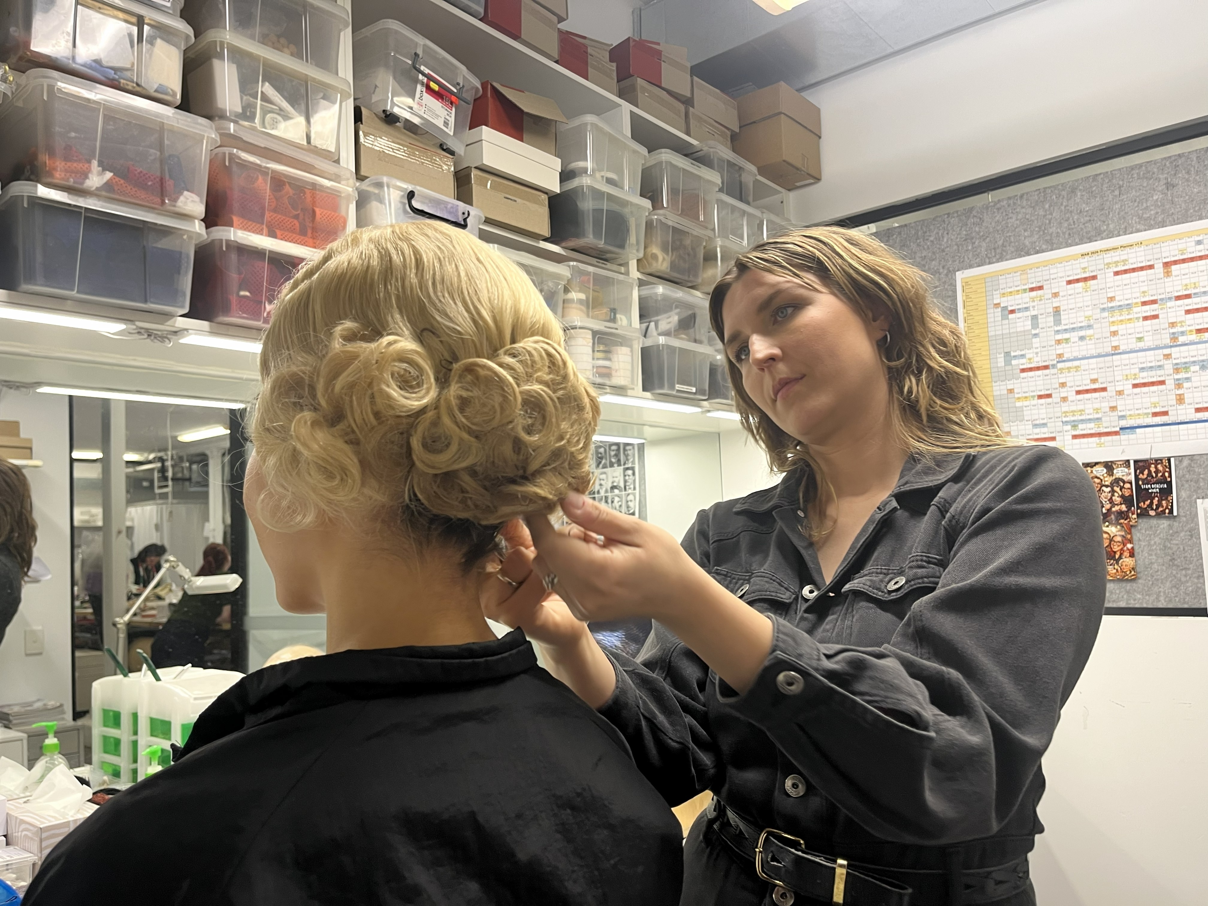 A woman adjusts a wig on a dancer's head as she sits in front of a mirror.