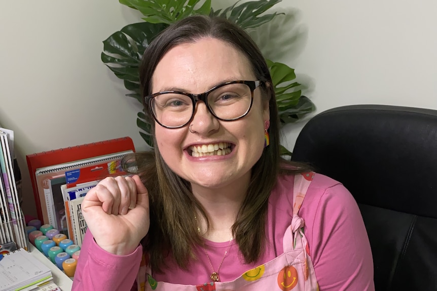 A woman in bright overalls sits at a desk and grins at the camera