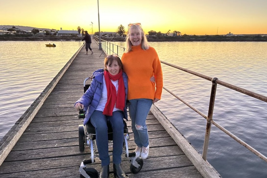 Two women stand on a pier at sunset. One woman sits in a wheelchair, their arms around each other