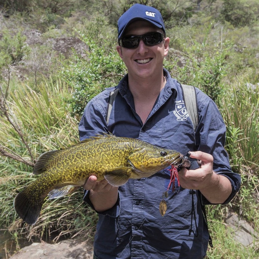 A man in a cap and sunnies smiling as he holds up a big river fish he has caught.