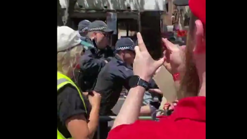 Police clash with a protester while a man films on his phone.