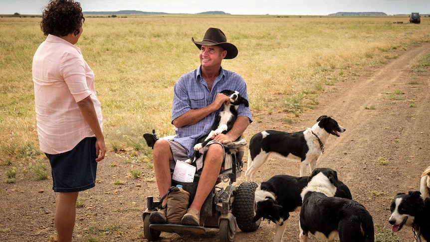 A man wearing an akubra and sitting in a motorised wheelchair chats to actress Deb Mailman surrounded by border collie puppies.