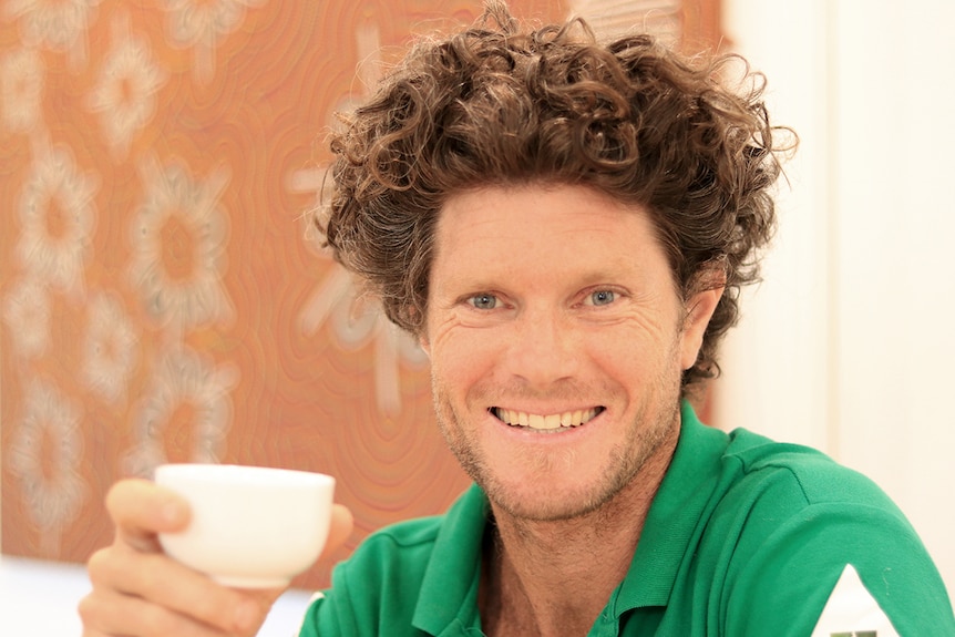 Mark Thirlwall smiles while holding a cup of tea, wearing a green polo shirt.