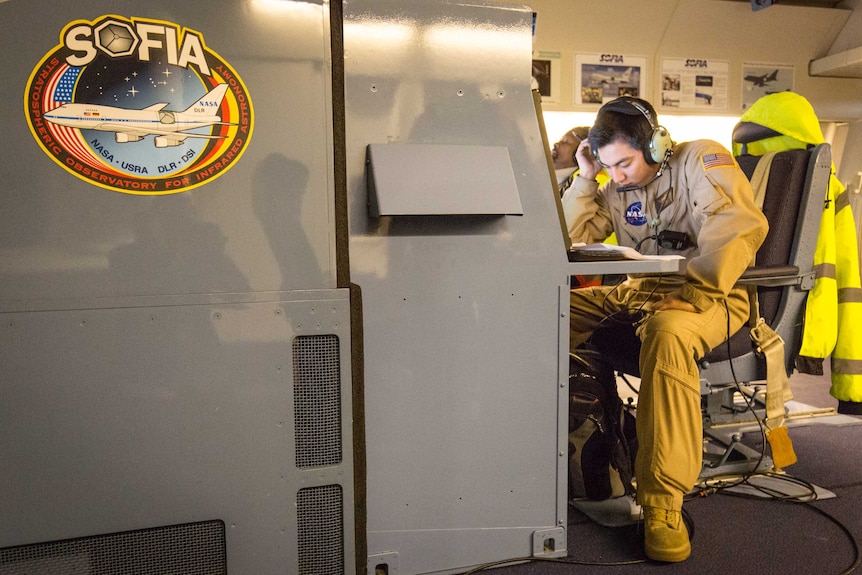 A man wearing a NASA jumpsuit and a radio headset sits hunched over paperwork on a huge computer console.