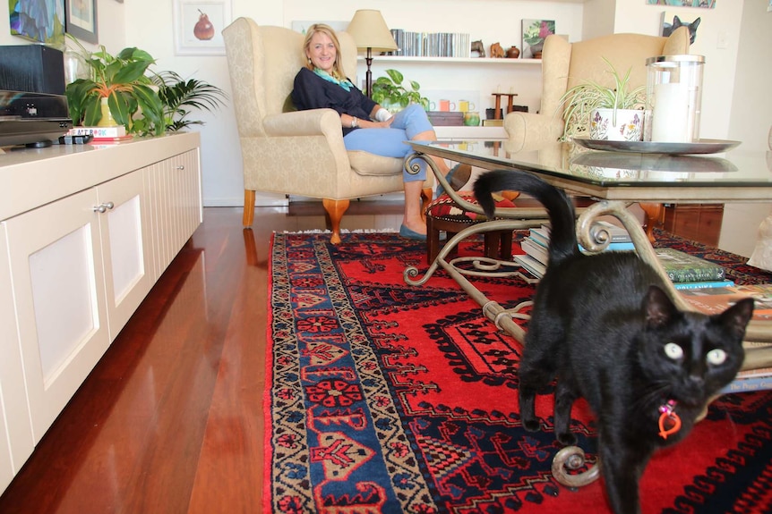 A woman sits in an armchair in her home with her arm bandaged as a black cat stands in the foreground.