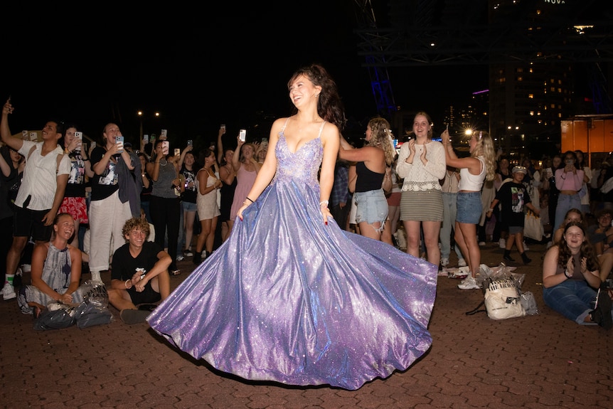 A woman in a ball gown twirls in front of smiling onlookers. 