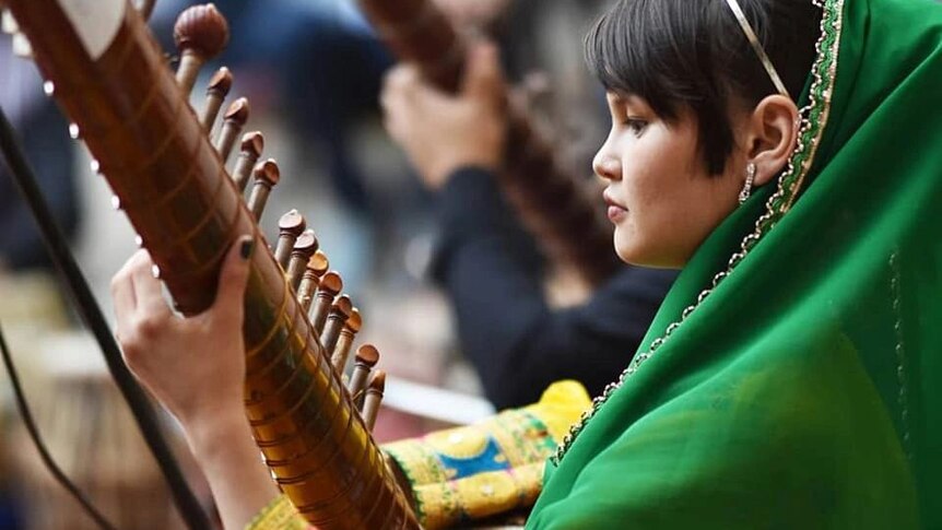 A member of the Zohra Orchestra wears a green headscarf and bright yellow top, playing an instrument while sitting on the floor.