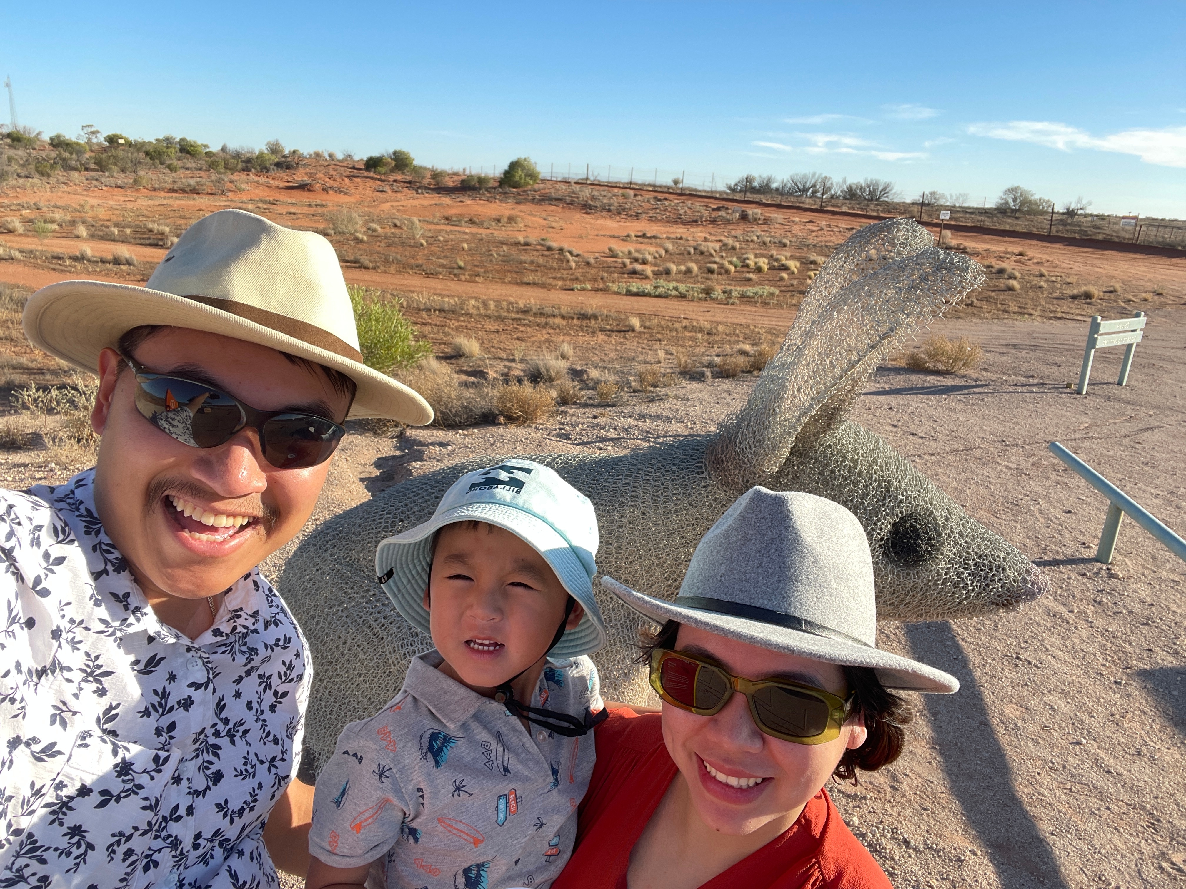 A woman, small child and man smile at the camera behind them is red dust. 