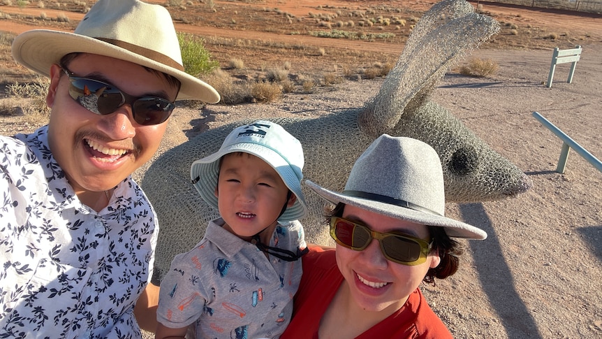 A woman, small child and man smile at the camera. Behind them is red dust and blue skies.All wear hats, adults sunnies.