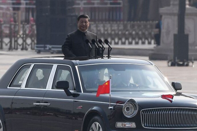 Xi Jinping standing from the sunroof of a black car with two small Chinese flags on the bonnet.