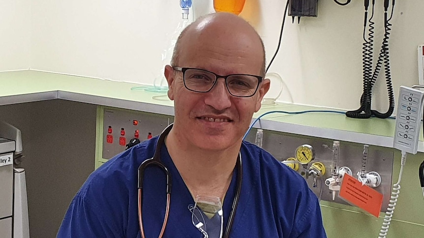 A man in a blue shirt and blue trousers sits on a hospital bed with a stethoscope around his neck and hospital equipment behind