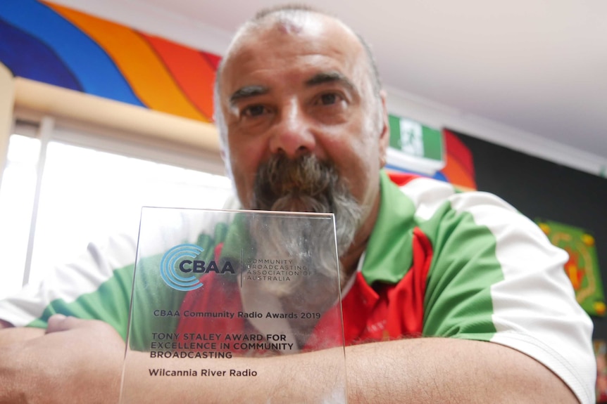 A perspex trophy is in focus, sitting on a bench, behind which a man in a beard looks at the camera, out of focus.