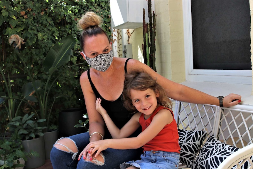 A mother wearing a face mask sits with her young daughter in a back yard posing for a photo.