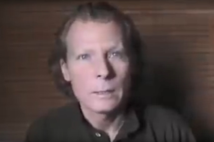 A man wearing a black shirt speaks to the camera in front of a brown backdrop.