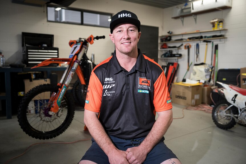 A smiling man sits in a garage in front of a dirt bike.