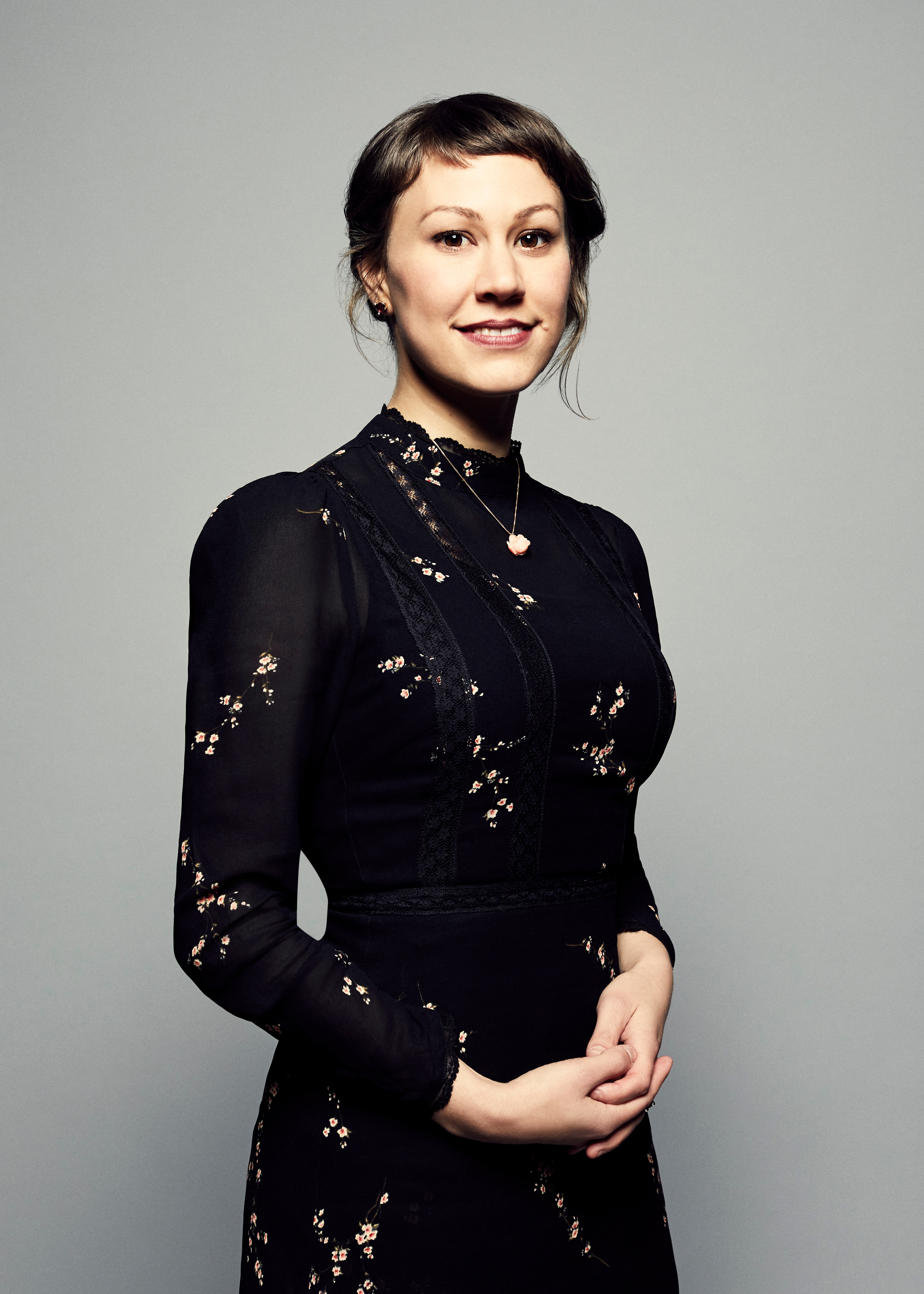 A white woman with brown hair pulled back with a fringe wears a floral black high-necked dress and smiles against grey backdrop