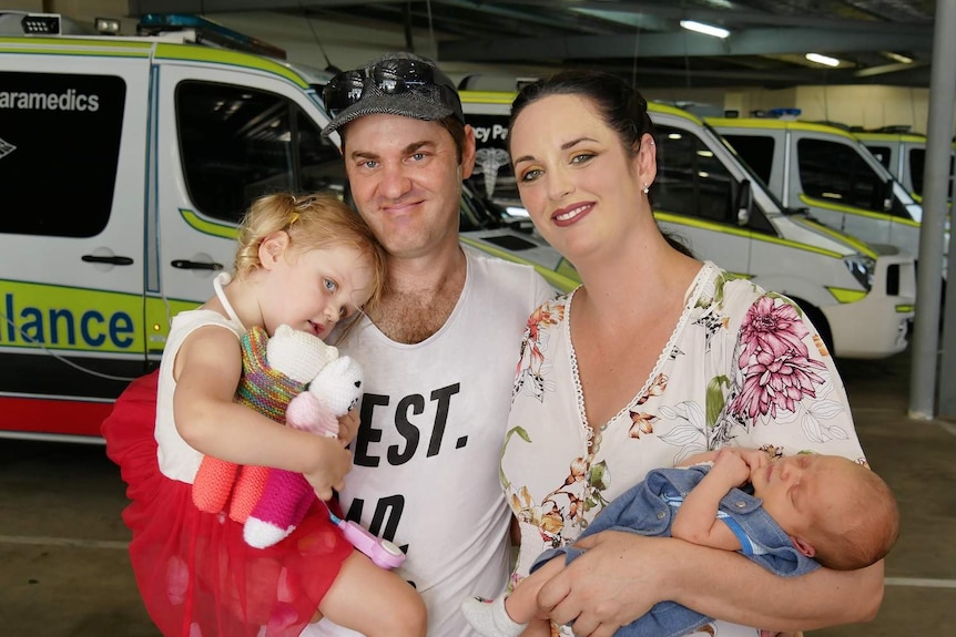 Carl, Alyssa-Marie Menzel with daughter Elyce and newborn Carlo together smiling at camera with ambulances in background.