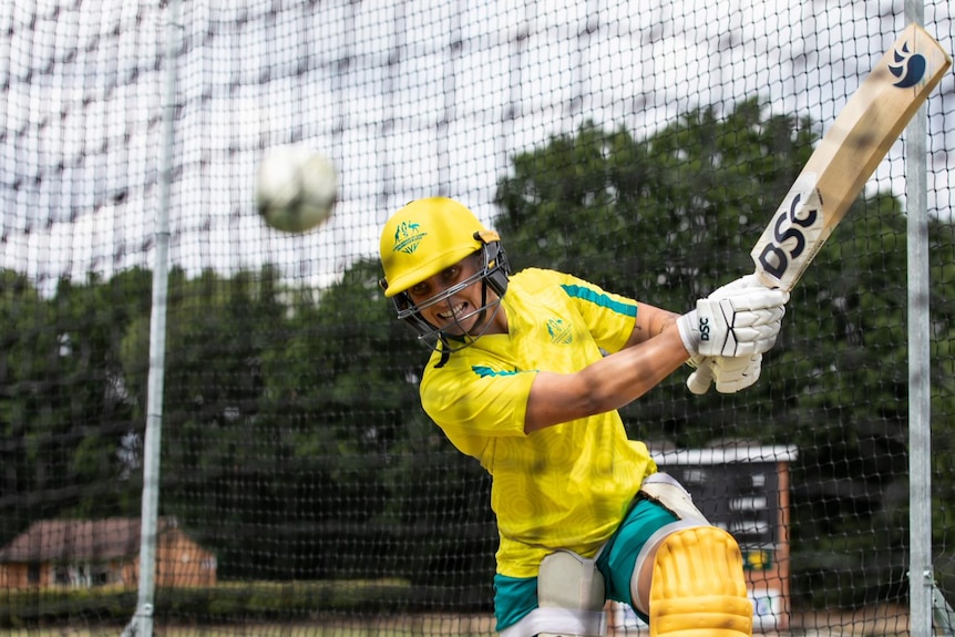 Ash Gardner hitting cricket ball in the nets. she wears her green and gold training clothes.