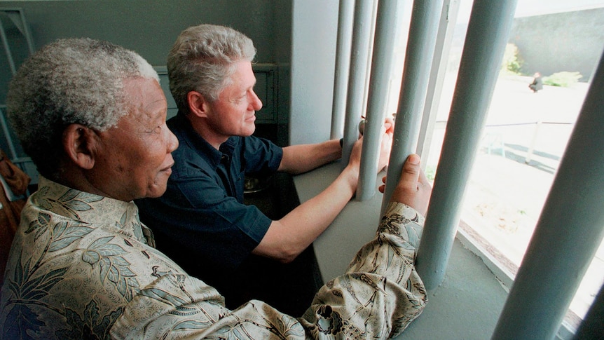 Nelson Mandela and Bill Clinton stand next to each other holding prison bars looking outside a prison window together
