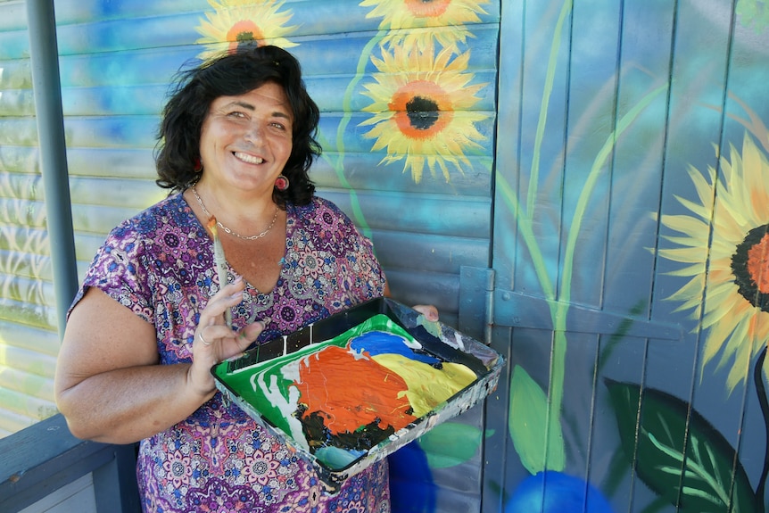 Woman wearing colourful dress sits next to painted sunflower mural, smiling and holding more paint.