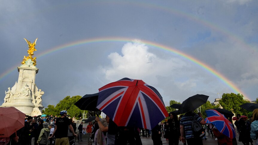 People with Union jack umbrellas look up to the sky with a rainbow outside Buckingham Palace. 