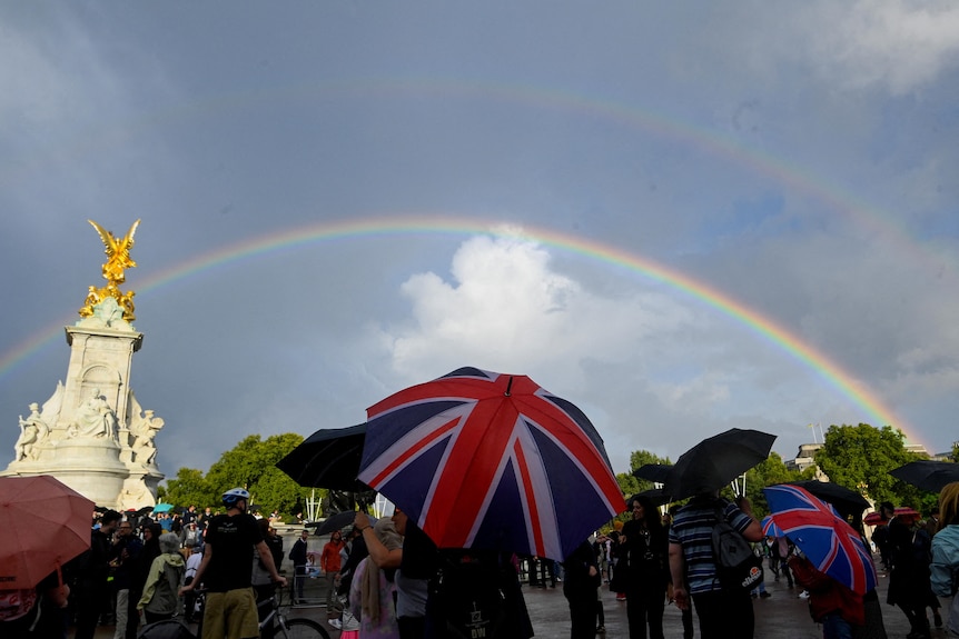People with Union jack umbrellas look up to the sky with a rainbow outside Buckingham Palace. 