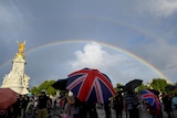 People with Union jack umbrellas look up to the sky with a rainbow outside Buckingham Palace. 