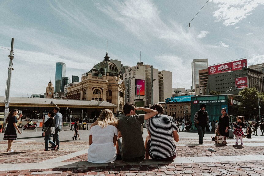 three young people, perhaps teenagers and recent high school leavers who are friends, sit together in Melbourne's Fed Square.
