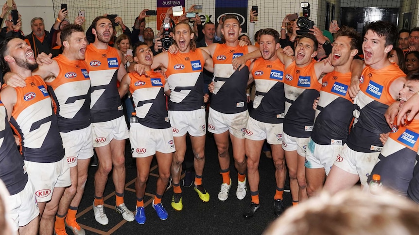 AFL players stand in a huddle in the dressing room singing their team song after victory